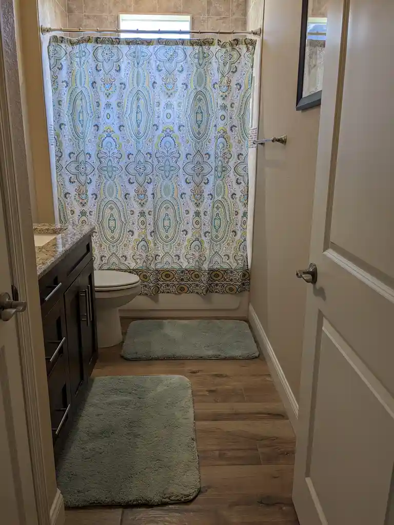 A bathroom with a wooden floor, two green rugs, a toilet, and a dark vanity with a granite countertop. The shower area is…