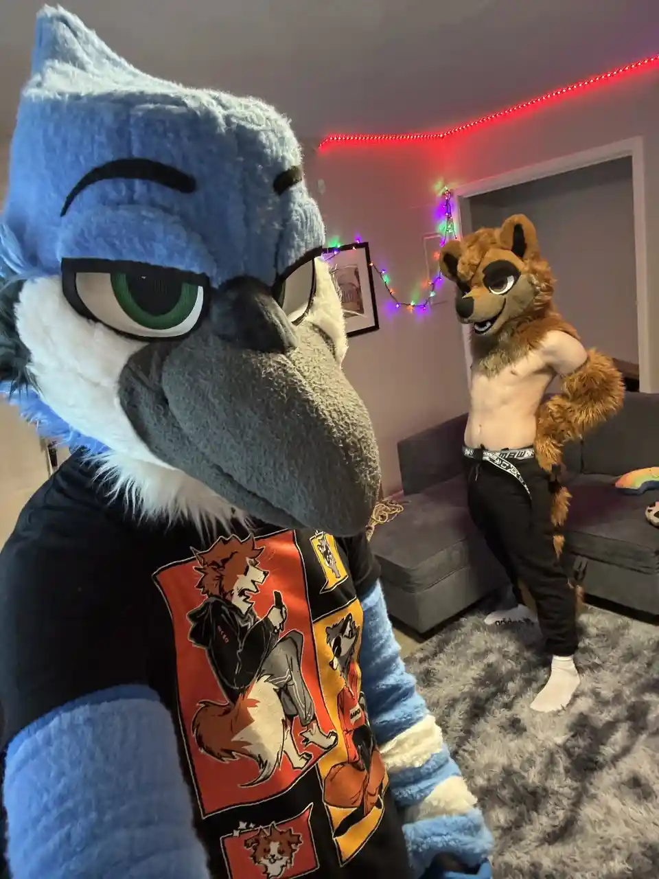 A person in a blue bird-like fursuit with glasses and a printed t-shirt looks sideways with a smug expression, while another…
