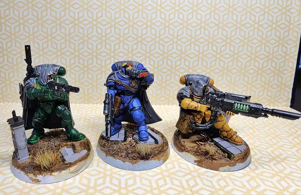Three painted miniature sci-fi soldiers in green, blue, and yellow armor stand in combat poses on textured bases against a…