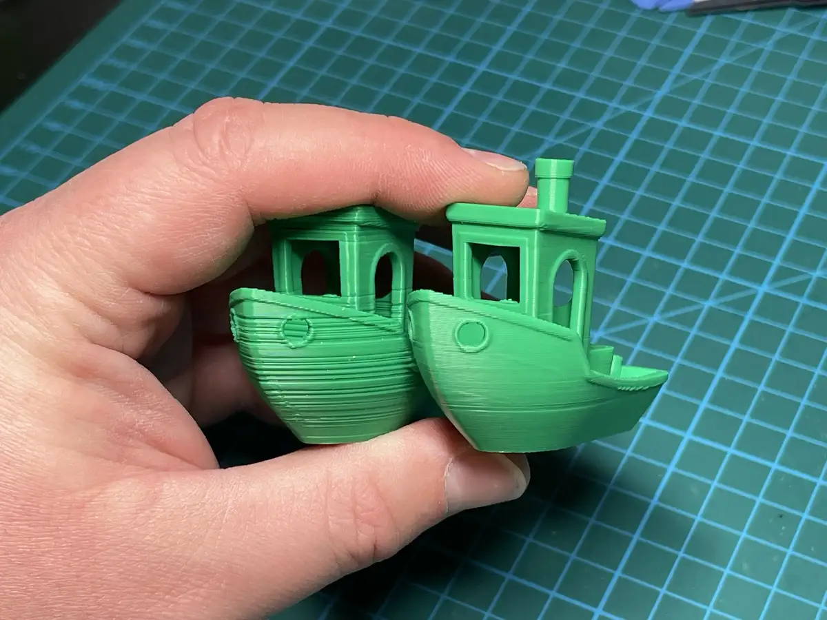A hand holds two small, identical green 3D-printed boat models side by side against a green grid cutting mat. The boats have…