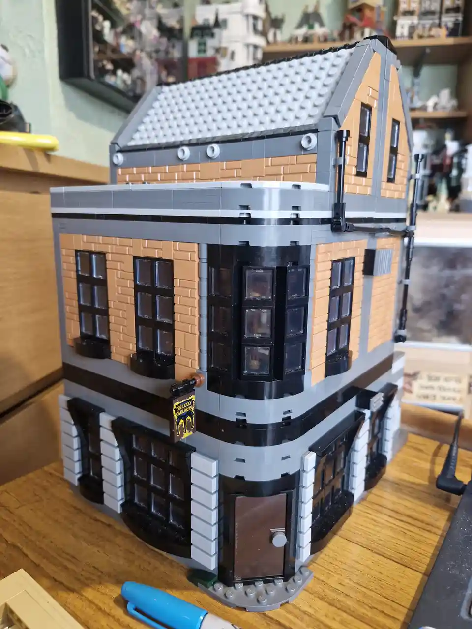 A detailed LEGO model of a two-story building with tan brick walls, black windows, and a gray roof, featuring a sign that…