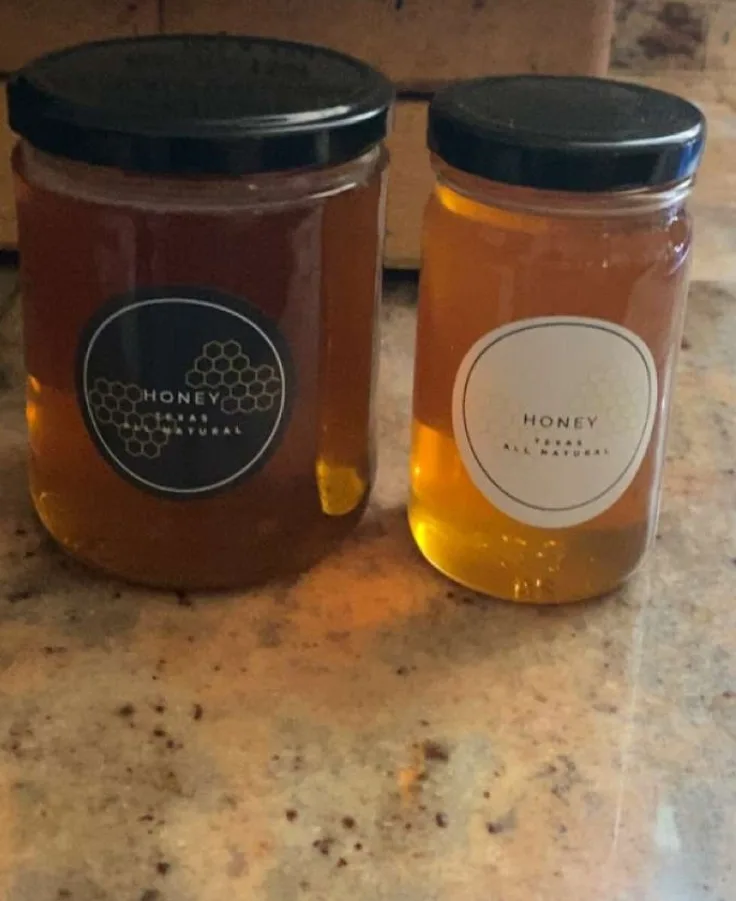 Two glass jars of honey sit side by side on a speckled countertop, each with a black lid and a label reading 'HONEY TEXAS…
