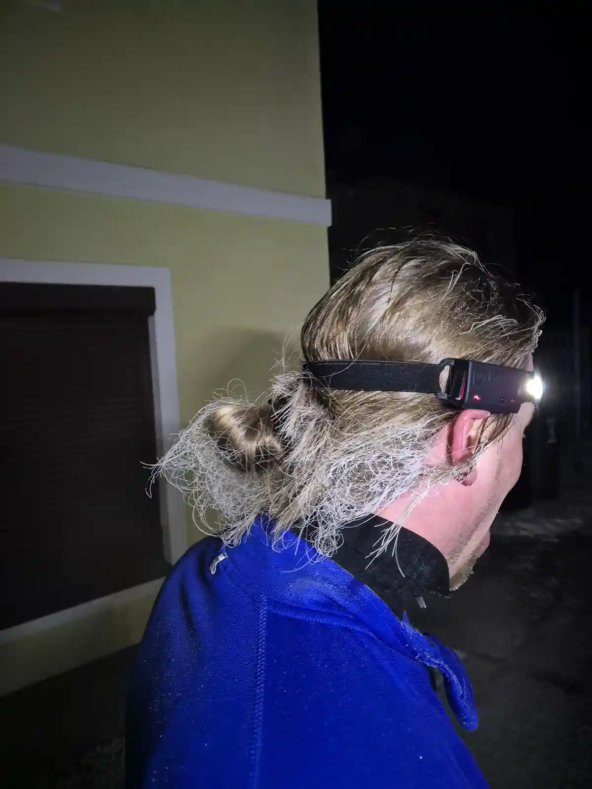 A person with blonde hair tied back is shown from the side at night, wearing a blue jacket and a headlamp that is turned on…