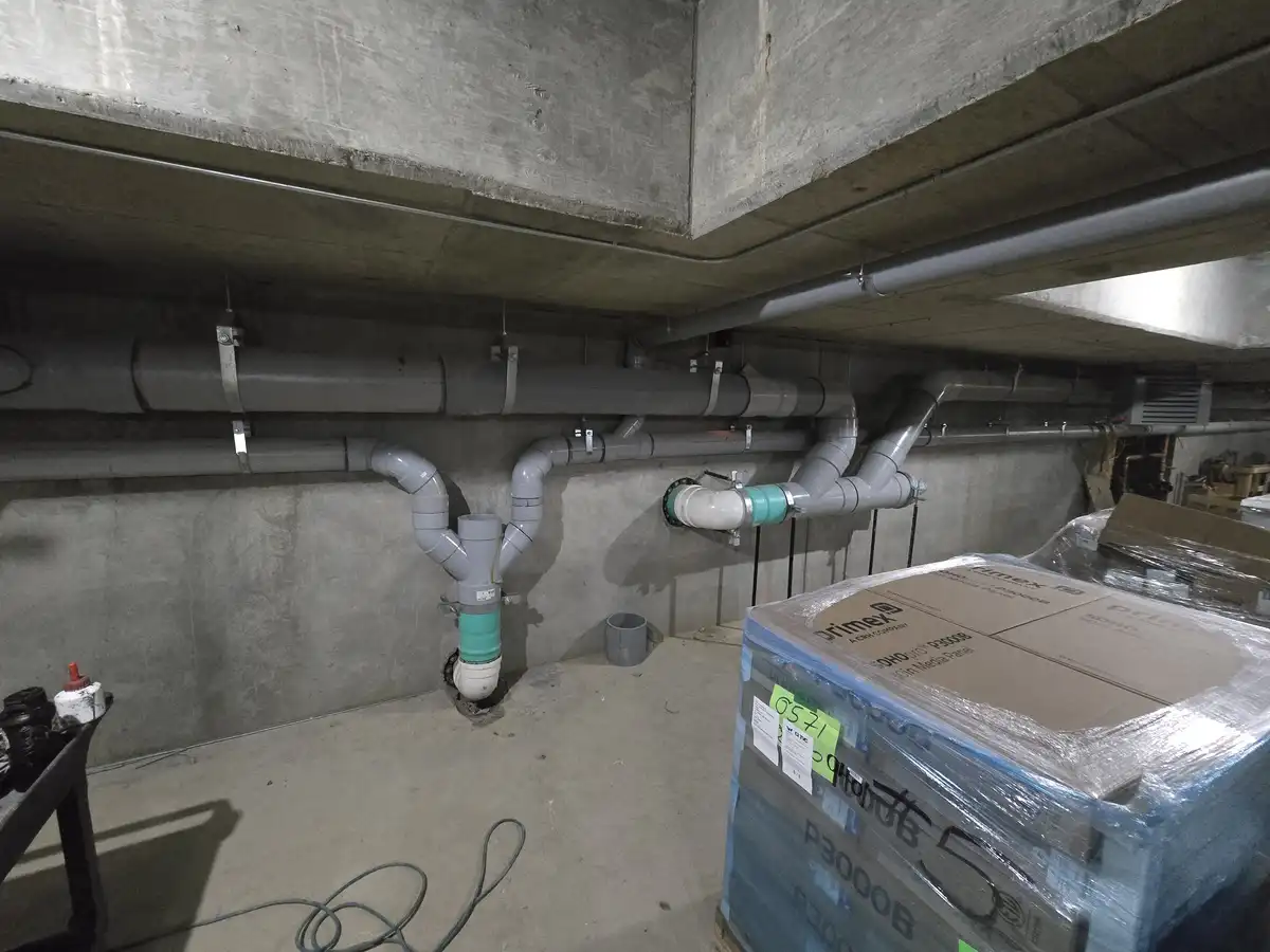 A dimly lit basement with exposed concrete walls and ceiling, featuring a complex network of gray PVC pipes running along…