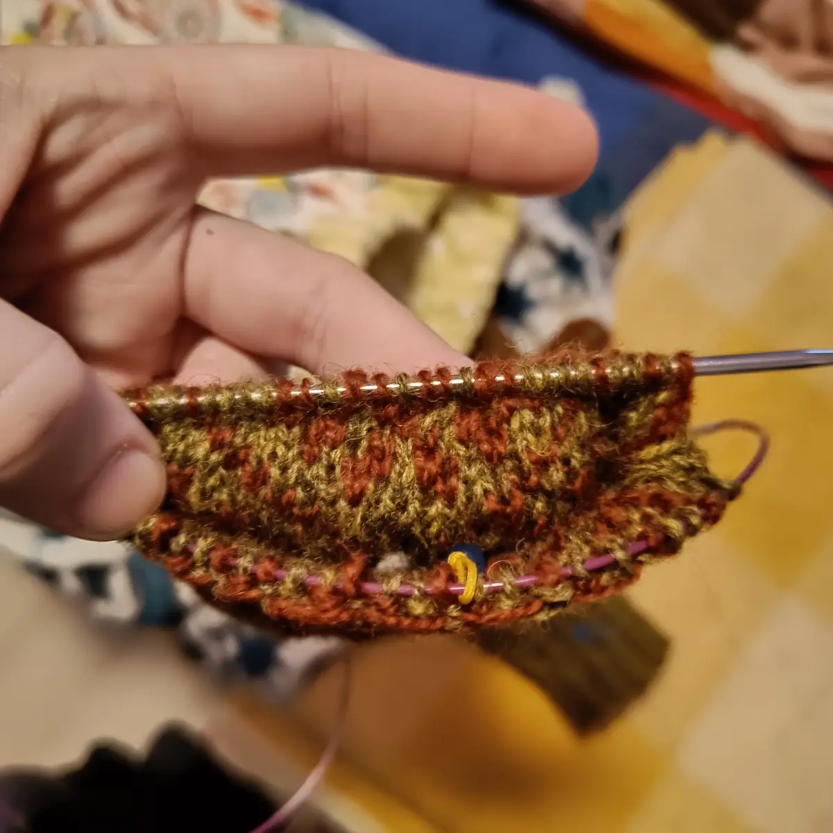 A close-up photo shows a hand holding a partially knitted piece with brown and green yarn on metal and pink knitting…