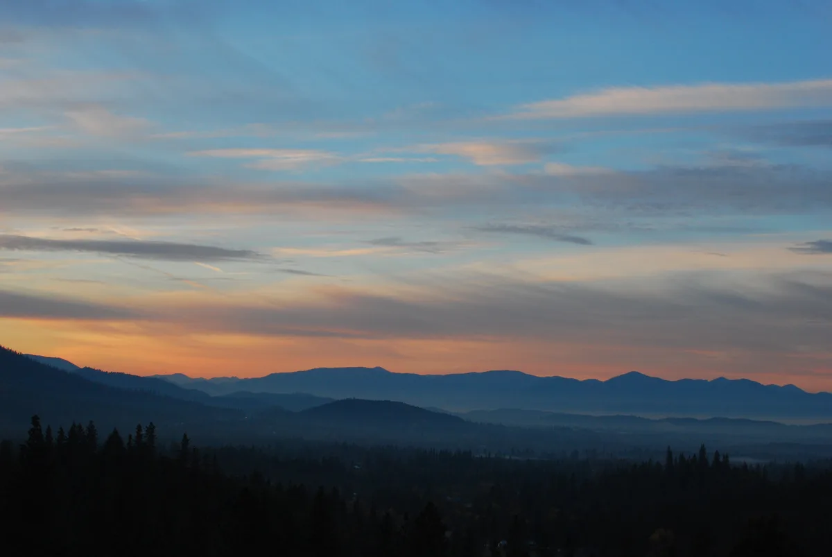 A serene landscape photo of layered mountain ranges silhouetted against a twilight sky with soft orange and blue hues. The…