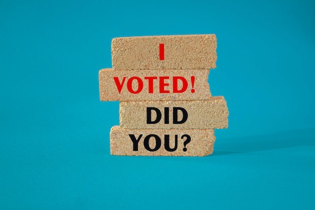 Four stacked cork blocks on a blue background, each bearing text: top block says 'I' in red, second says 'VOTED!' in red…