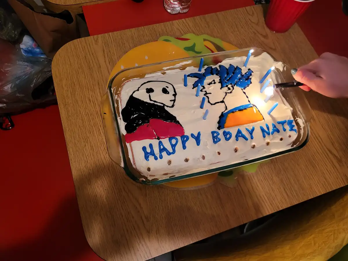 A homemade birthday cake in a glass baking dish features a cartoon drawing of a panda and a person with blue hair, both…