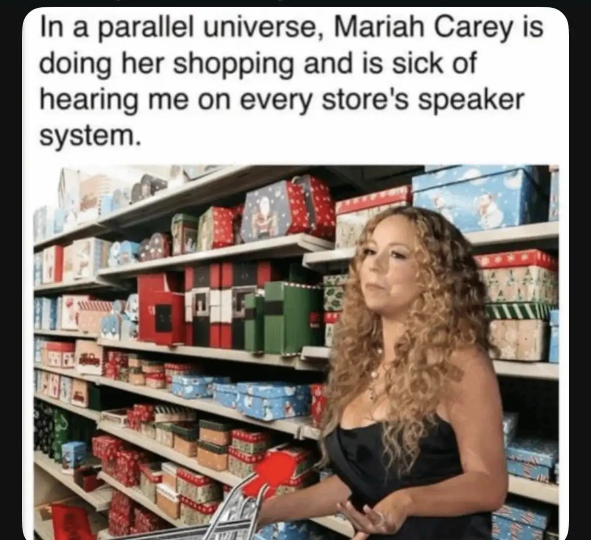 A woman with long curly hair, identified as Mariah Carey, stands in a store aisle filled with colorful holiday gift boxes…