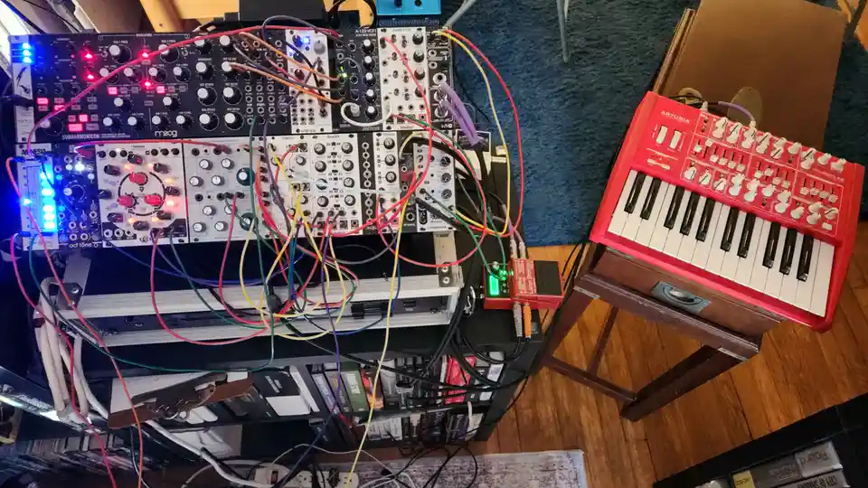 An overhead photo shows a cluttered music studio setup with a red Arturia Minibrute R4 synthesizer on a stand to the right…
