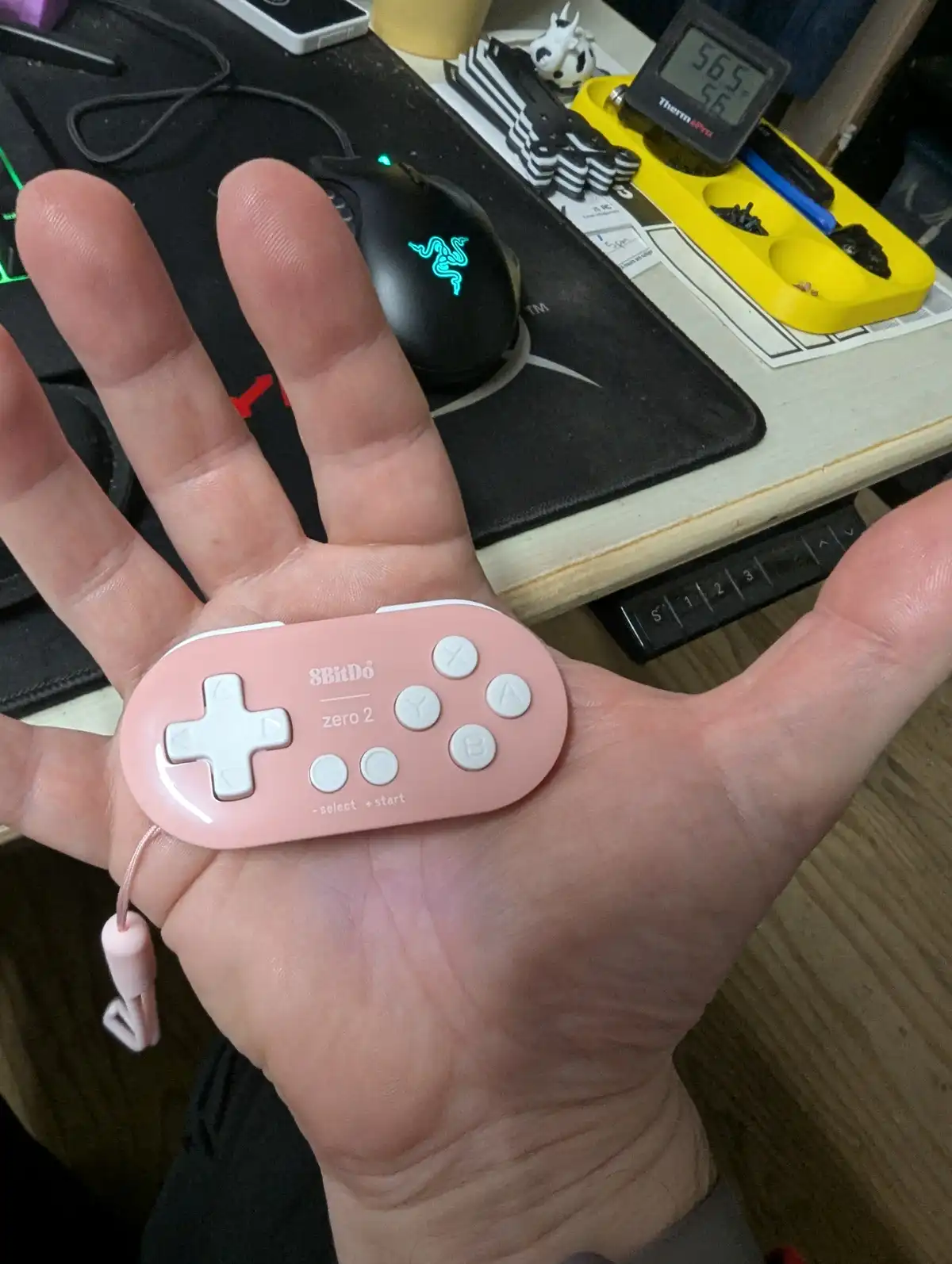 A person holds a small, pink 8BitDo zero 2 game controller in their open palm, showcasing its compact size. The background…
