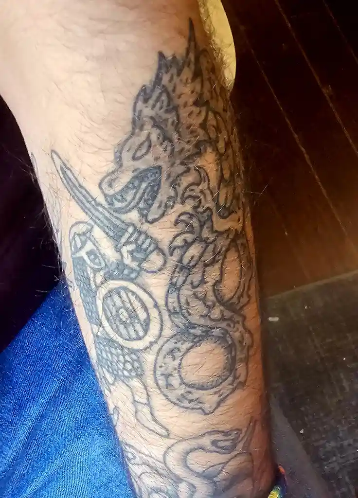 A close-up photo shows a person's forearm with a large, intricate black-and-gray tattoo featuring a stylized dragon or…