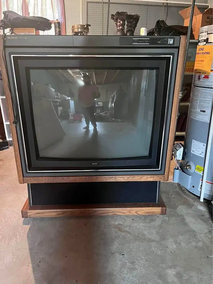 A large, old-style CRT television with a wooden frame sits in a cluttered garage or storage room, its screen reflecting the…