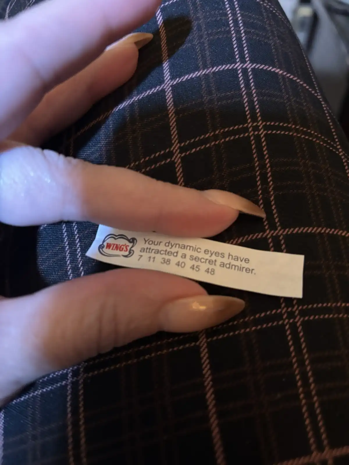 A hand with long, light-brown fingernails holds a small white fortune cookie slip from 'WINGS' against a dark plaid fabric…