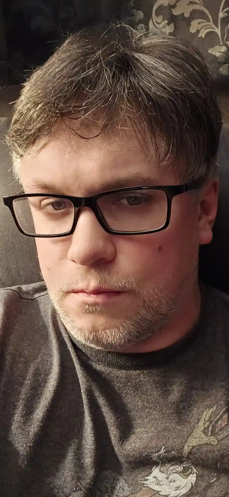 A close-up photo of a man with short brown and gray hair, wearing black-rimmed glasses and a gray t-shirt, looking directly…