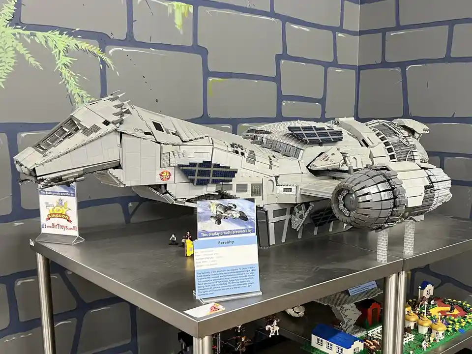A large, detailed LEGO model of the spaceship Serenity from the TV show 'Firefly' is displayed on a metal shelf. The ship is…