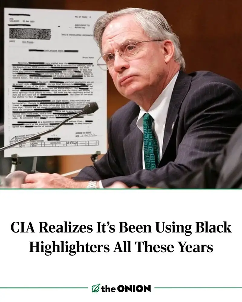 A man in a suit and glasses sits at a table with a microphone, looking concerned. Behind him is a document with redacted…