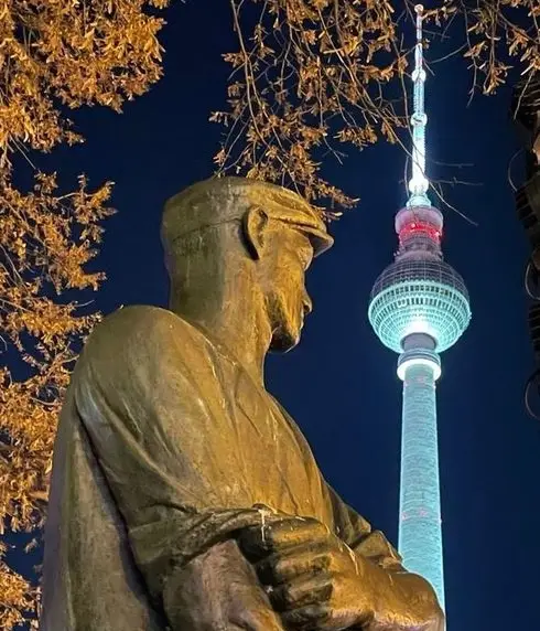 A bronze statue of a man in a cap, viewed from the side, stands in the foreground at night. Behind him, the illuminated…