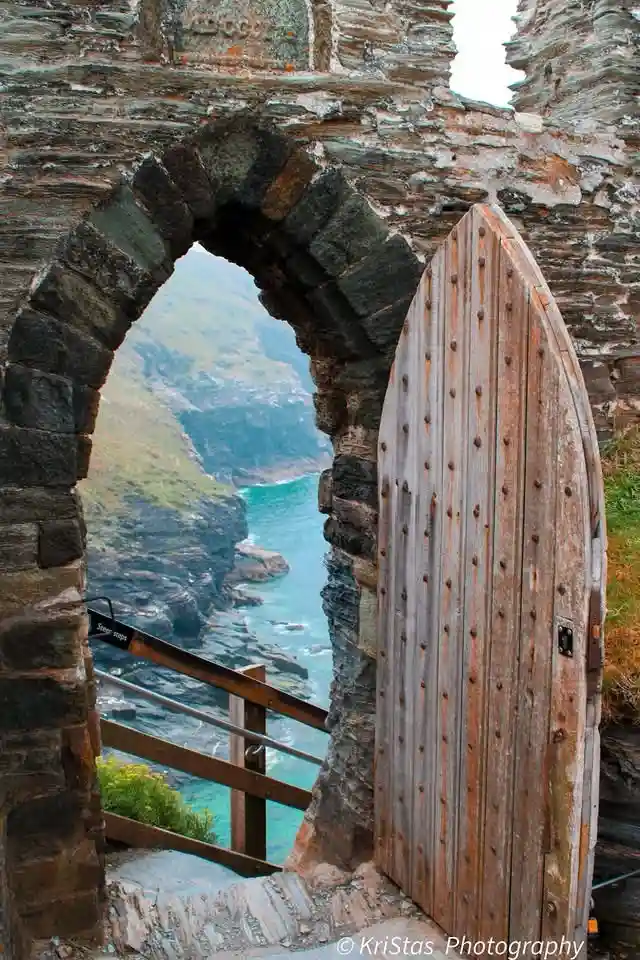 A weathered wooden door is ajar within a stone archway of a coastal ruin, revealing a stunning view of turquoise ocean and…