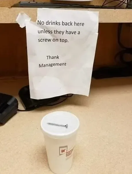 A white styrofoam cup with a metal screw inserted into its lid sits on a beige desk surface. Taped to the desk above it is a…
