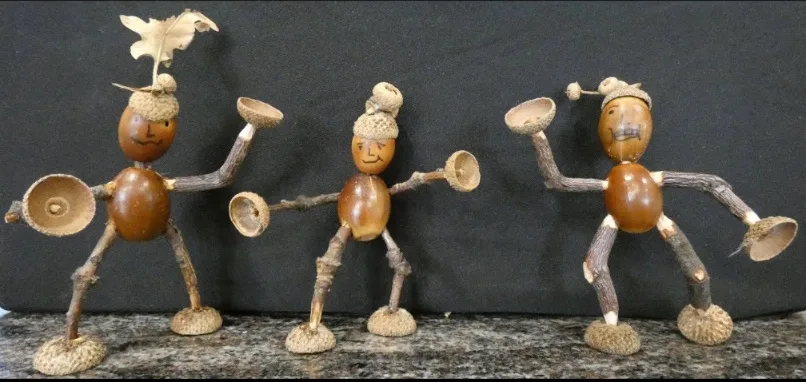 Three handmade figurines made from acorns, twigs, and oak leaves are posed in a line, each holding acorn caps as if dancing…