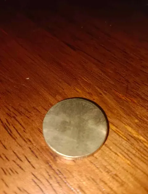 A close-up photo shows a plain, round, silver-colored coin resting on a dark wooden surface with visible grain. The overlaid…