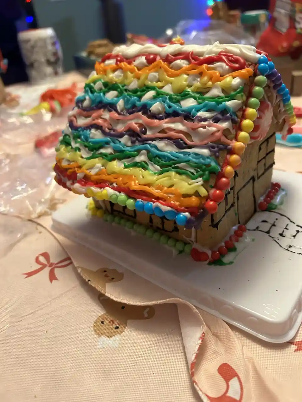 A gingerbread house decorated with multiple wavy layers of rainbow-colored icing and colorful candy beads along the edges…