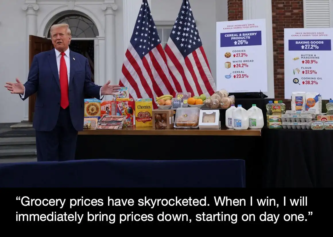 A man in a blue suit and red tie stands behind a table laden with cereal boxes, bread, milk, eggs, and other groceries. Two…