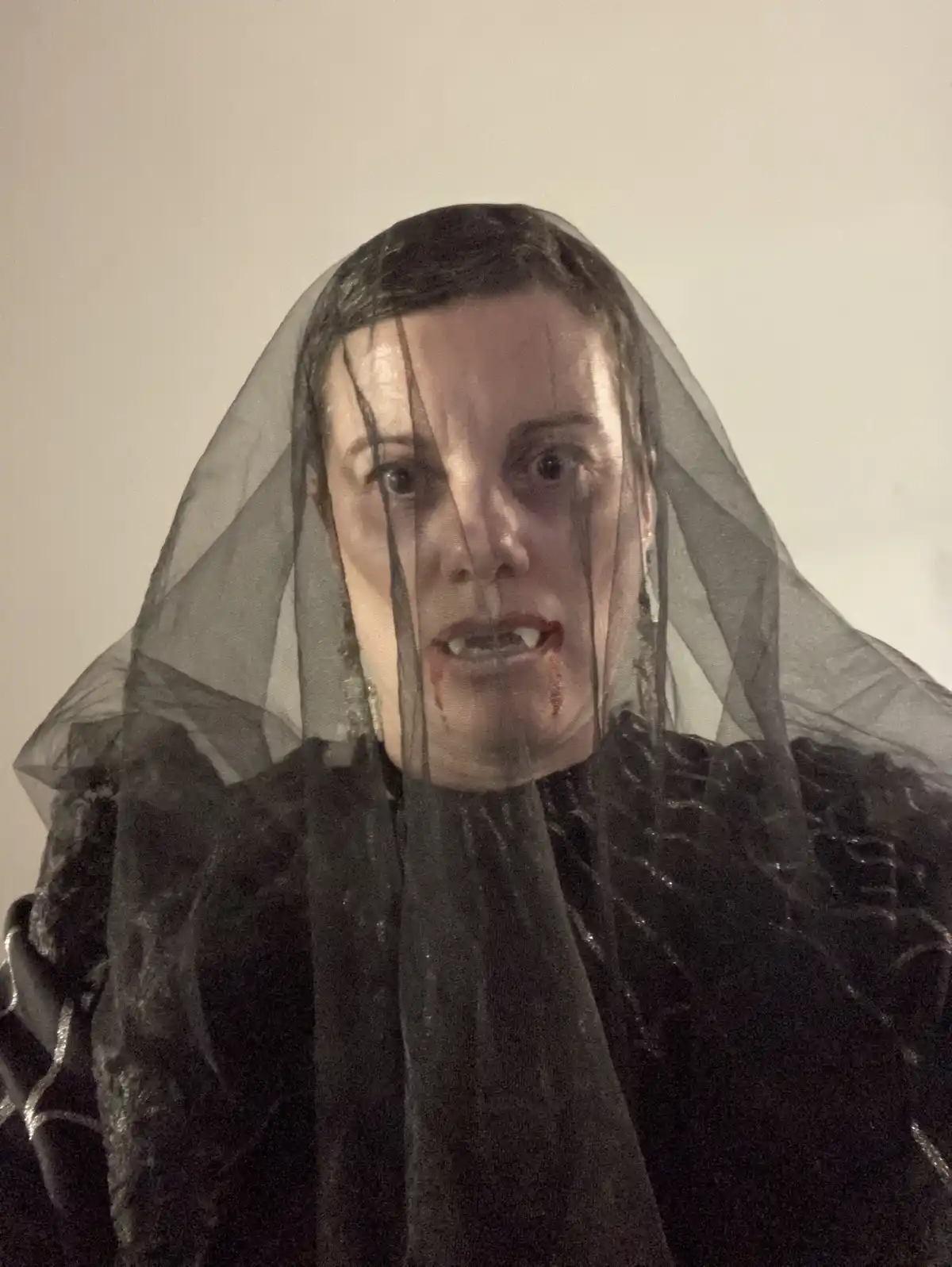 A person dressed in a black, ruffled costume with a sheer black veil over their head, showing a face with fake fangs and…