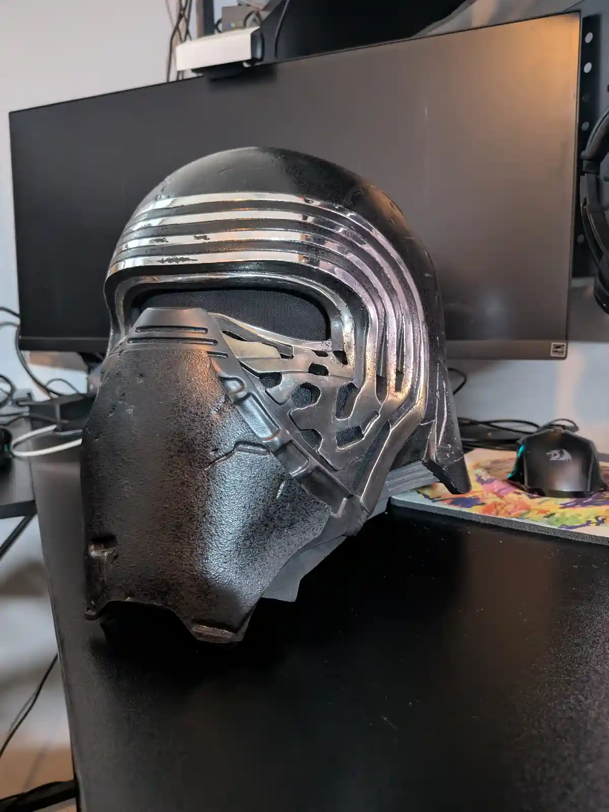 A black, glossy Kylo Ren-style helmet from Star Wars sits on a black desk in front of a monitor, with visible wear and…