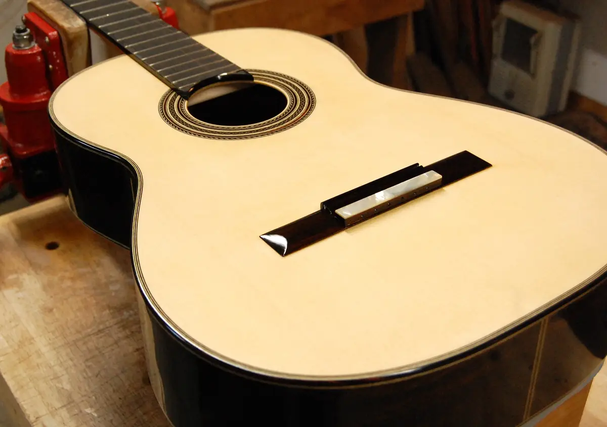 A close-up photo of a light-wood acoustic guitar with a dark, glossy finish, resting on a wooden workbench. The guitar's…