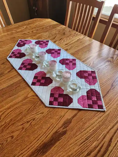 A wooden dining table features a handmade quilted table runner with pink and magenta heart patterns. Several small candles…