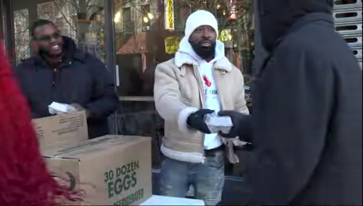 A man in a beige winter coat and white beanie hands a small box to another person wearing a black hooded jacket. Behind him…
