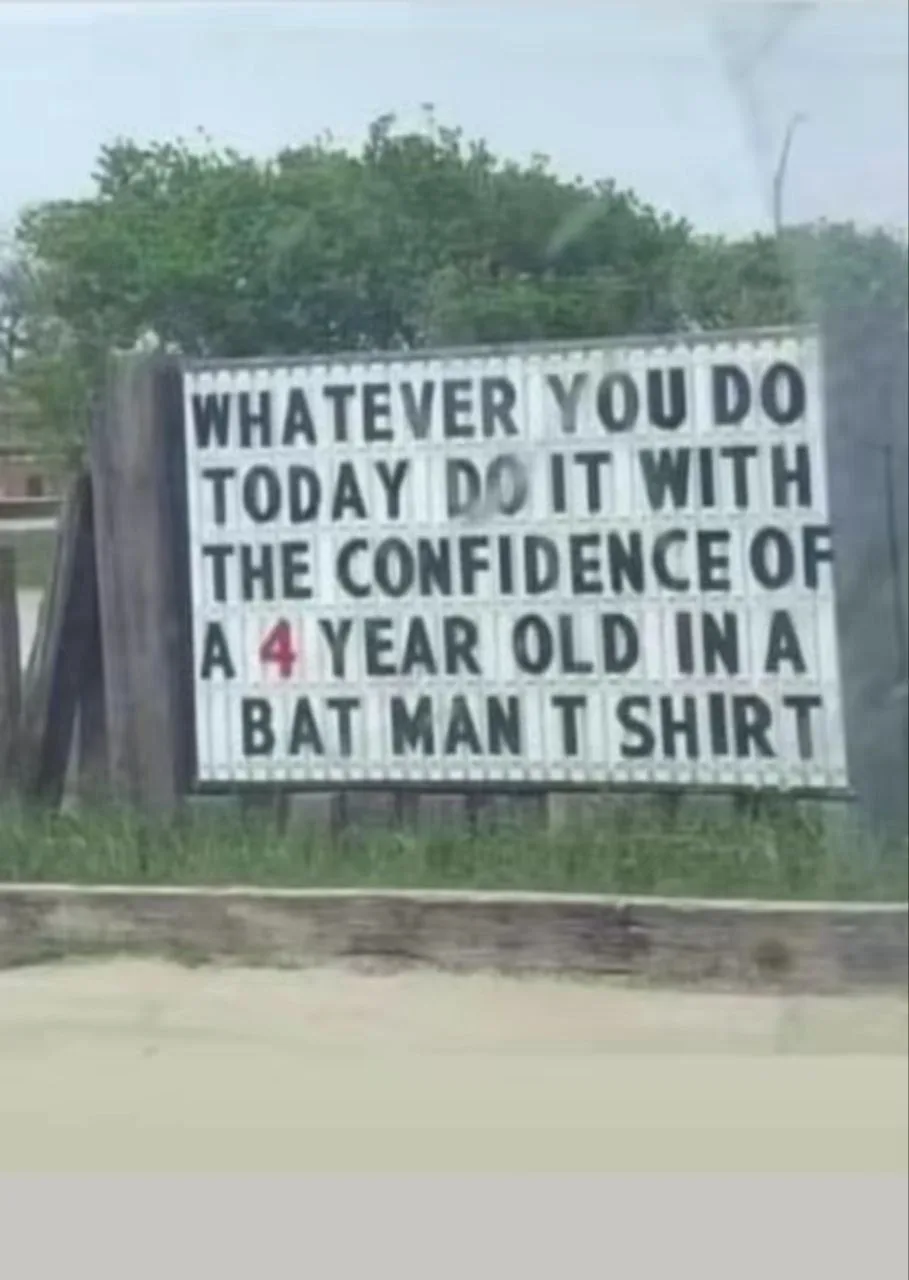 A roadside sign with black block letters on a white background reads: 'WHATEVER YOU DO TODAY DO IT WITH THE CONFIDENCE OF A…