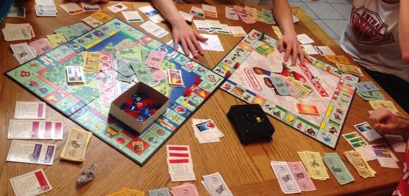 Two Monopoly game boards are laid out side-by-side on a wooden table, surrounded by scattered cards, money, and game pieces…