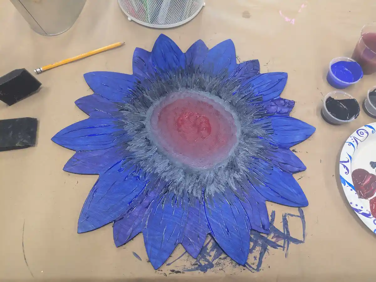 A hand-painted sunflower craft with blue petals and a red center sits on a brown paper-covered table, surrounded by art…