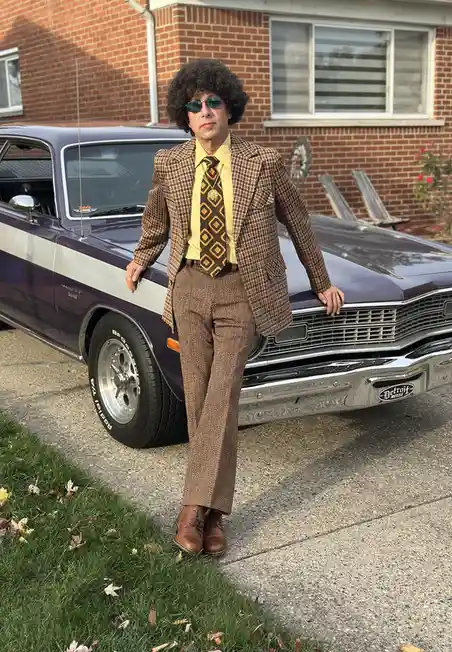 A man with a large curly afro and sunglasses poses confidently in front of a purple and white classic American muscle car…