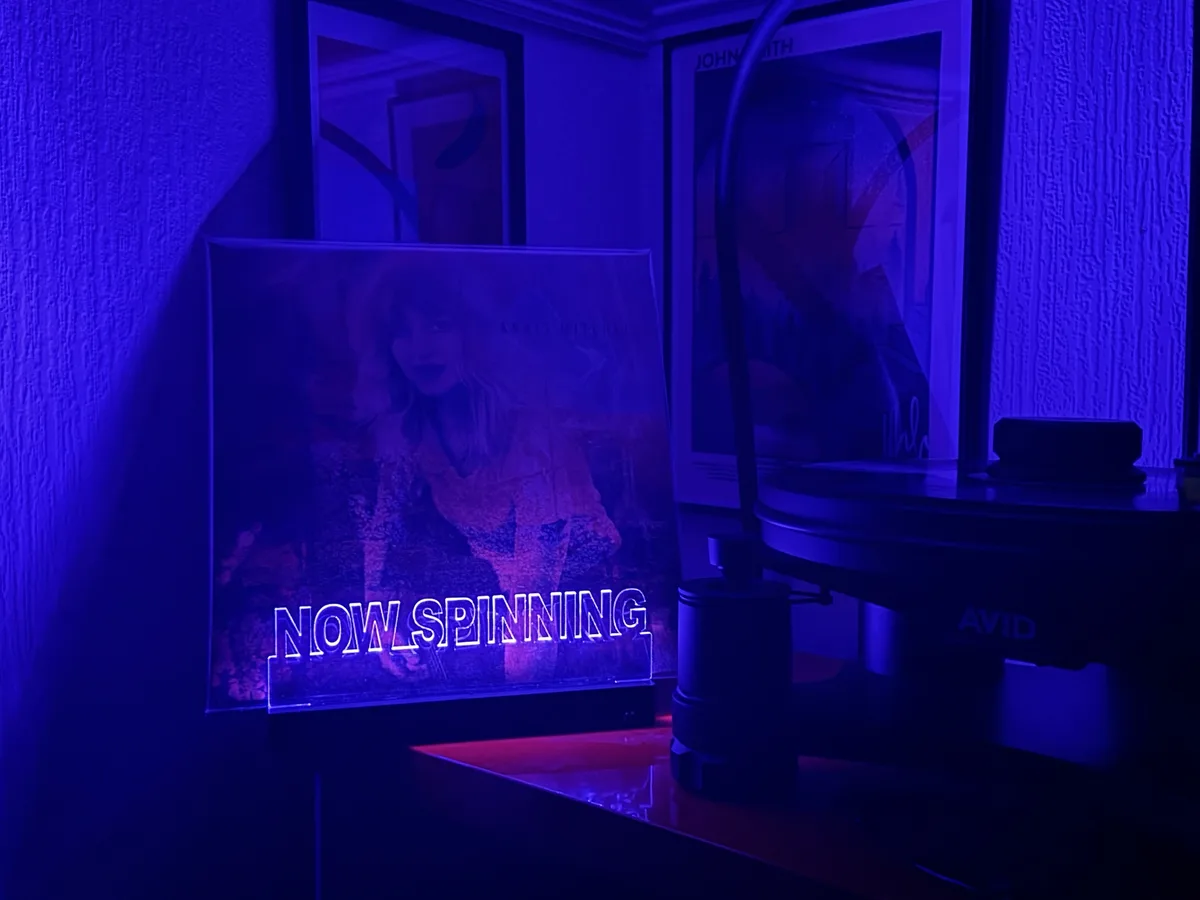 A dimly lit room bathed in blue light features a vinyl record player with an 'AVID' brand turntable. Next to it, a glowing…