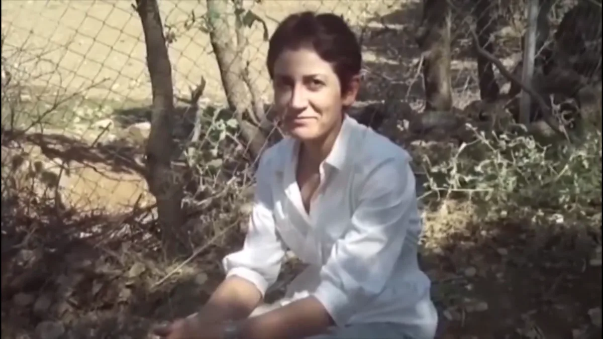 A woman with short dark hair wearing a white button-up shirt is crouching outdoors in front of a chain-link fence and dry…