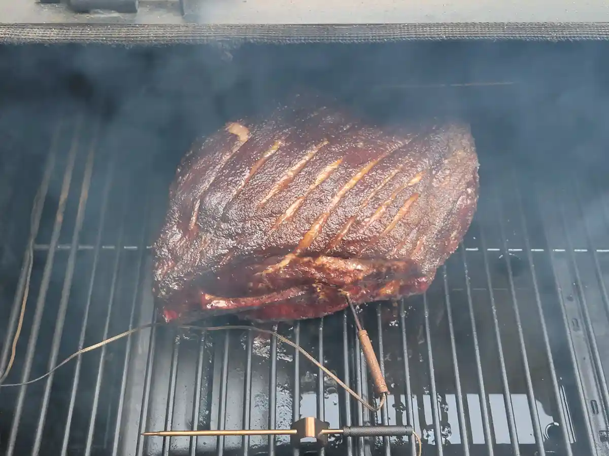 A large, heavily smoked piece of meat with diagonal scoring sits on a grill grate, surrounded by smoke, with a thermometer…