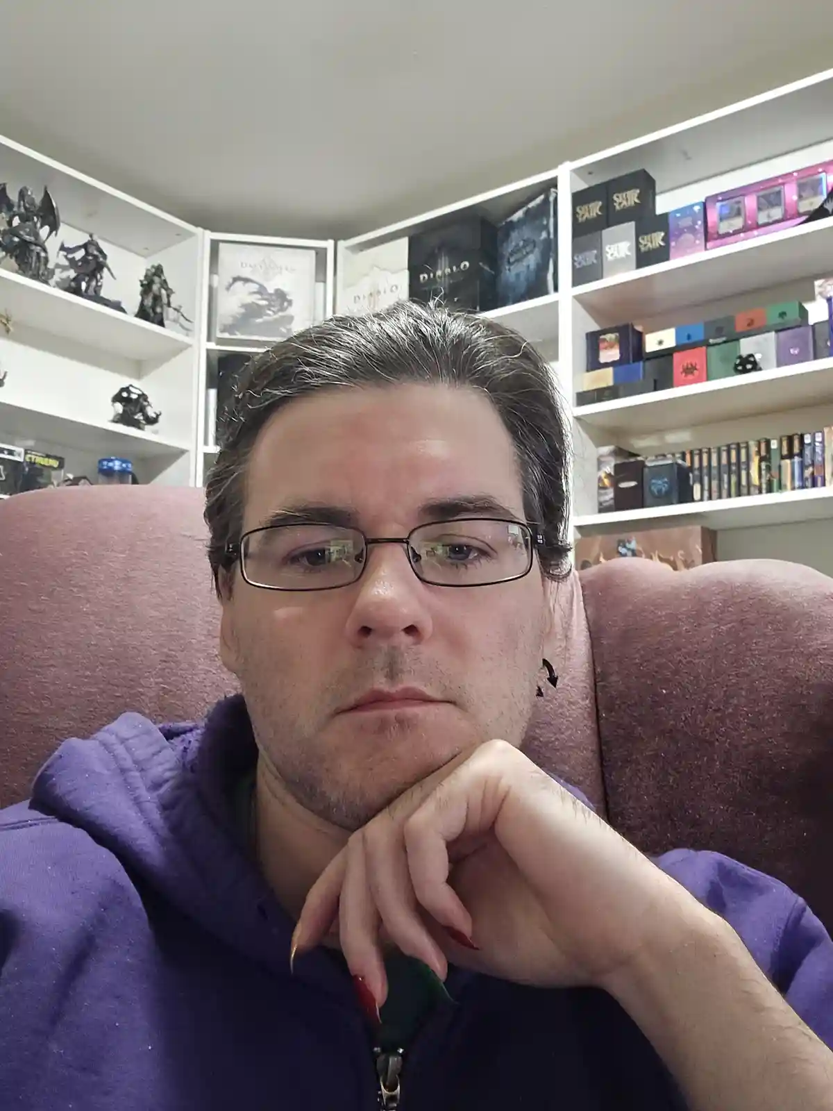 A man with glasses and a purple hoodie sits in a maroon armchair, resting his chin on his hand with a pensive expression…