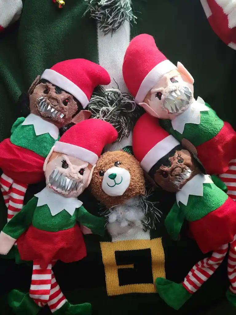 Four small, traditionally dressed Christmas elves with red hats and green outfits are arranged around a plush teddy bear on…