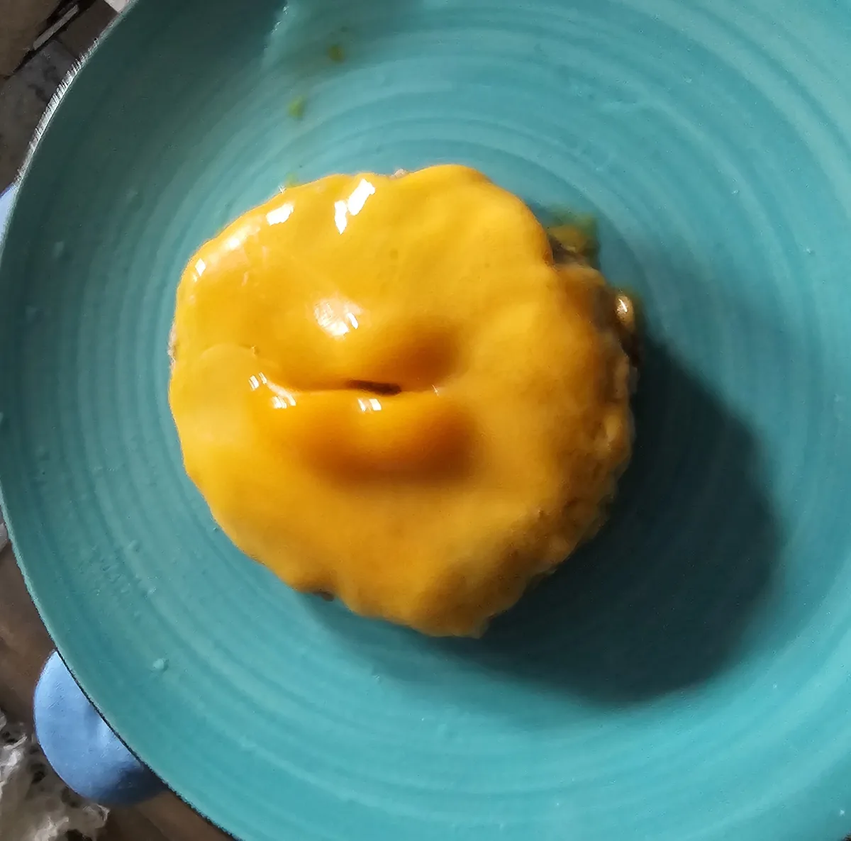 A close-up photo shows a round, yellow, cheese-covered food item on a teal plate with concentric ridges. The cheese has a…