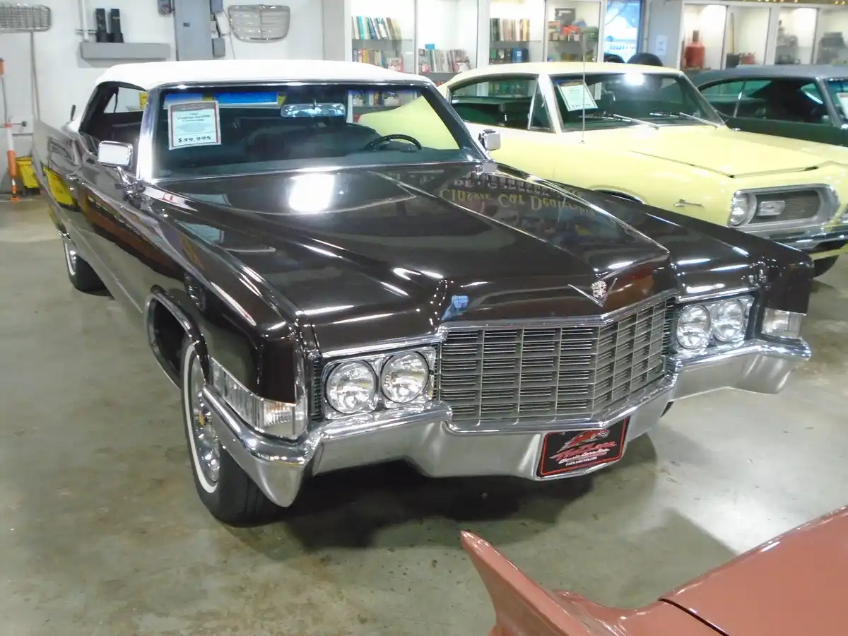 A shiny black 1960s Cadillac DeVille convertible is displayed in a classic car showroom, with a price tag of $39,995 visible…
