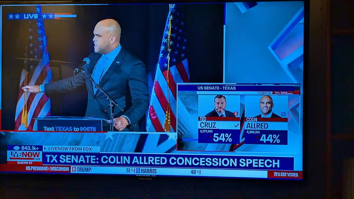 A television screen shows Colin Allred, a bald man in a suit, giving a concession speech at a podium with American flags…