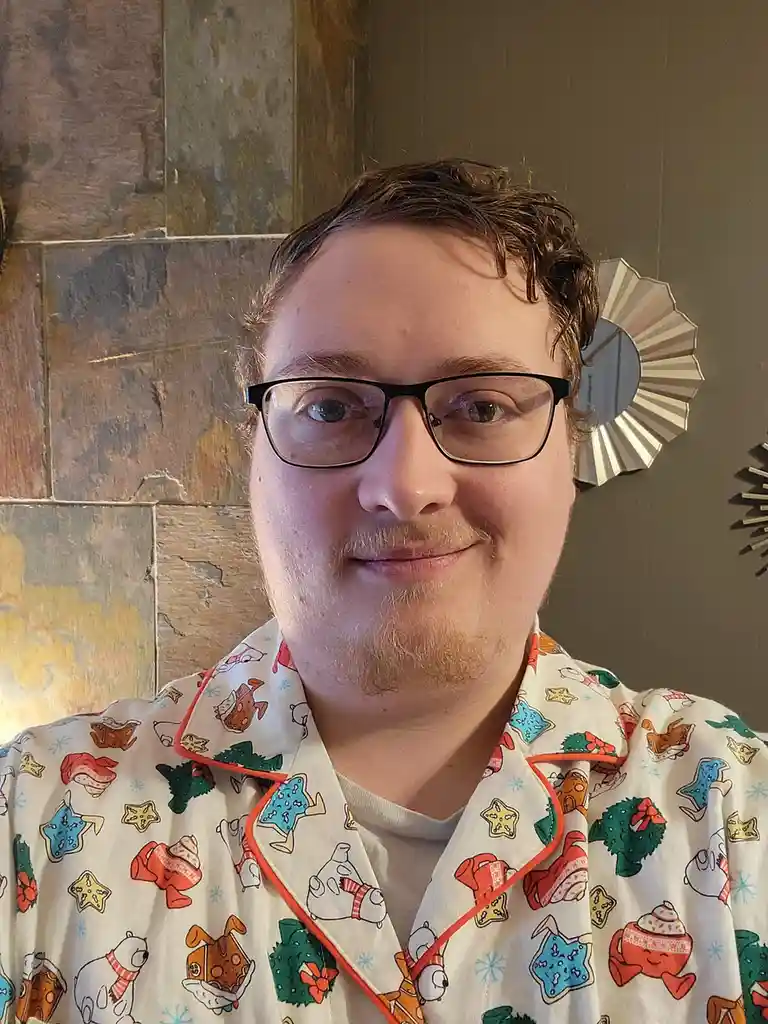 A person with glasses and a light beard wears festive, cartoon-themed pajamas featuring polar bears, gingerbread men, and…