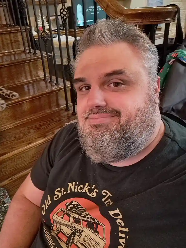 A man with a gray beard and hair wears a black t-shirt with a graphic of a van labeled 'Old St. Nick's Tree Delivery' and a…