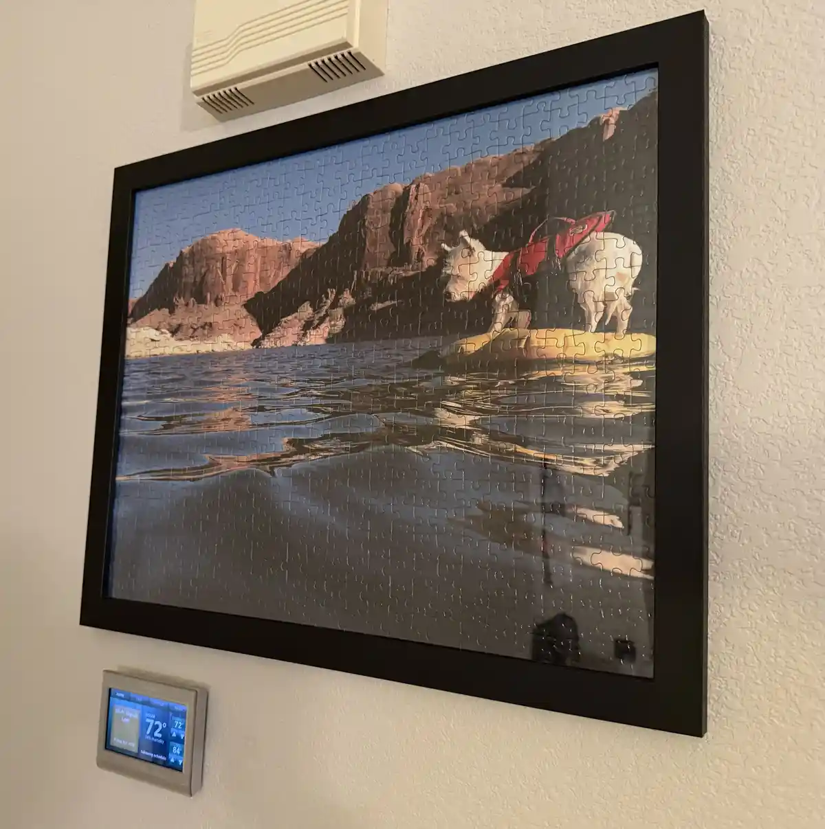 A framed puzzle depicting a white horse wearing a red vest standing on a yellow raft in water with rocky cliffs in the…