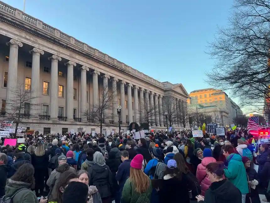 A large crowd of protesters gathers in front of a grand neoclassical building with tall columns under a clear blue sky. Many…