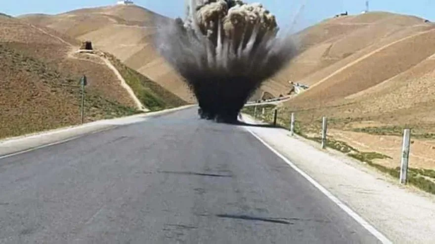A wide asphalt road stretches through a dry, hilly landscape under a clear blue sky. At the center of the road, a massive…